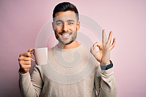 Young handsome man drinking a cup of hot coffee over pink isolated background doing ok sign with fingers, excellent symbol