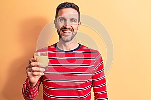 Young handsome man drinking glass of healthy orange juice over isolated yellow background looking positive and happy standing and