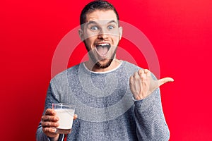 Young handsome man drinking glass of milk pointing thumb up to the side smiling happy with open mouth