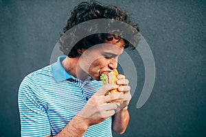 Young handsome man eating a healthy burger. Hungry man in a fast food restaurant eating a hamburger outdoors. Man with curly hair