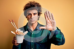 Young handsome man eating noodles standing over isolated yellow background with open hand doing stop sign with serious and