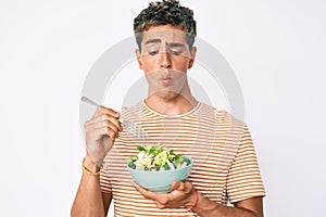 Young handsome man eating salad making fish face with mouth and squinting eyes, crazy and comical