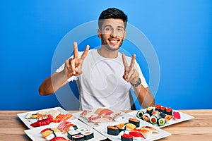 Young handsome man eating sushi sitting on the table smiling looking to the camera showing fingers doing victory sign