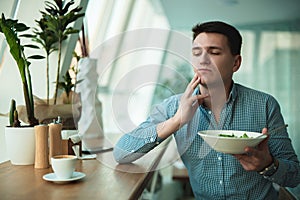 Young handsome man felt toothpain while eating salad and drinking coffee for lunch during break at cafe near office