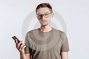 Young handsome man with glasses holding smartphone. Student, worker looks data information, posing for photo isolated on gray