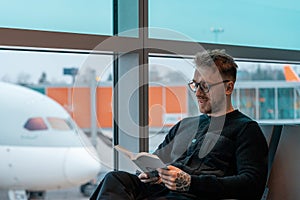 Young handsome man with glasses reading a book while waiting for boarding at air jet in airport.