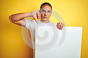 Young handsome man holding advertising banner over yellow isolated background with angry face, negative sign showing dislike with