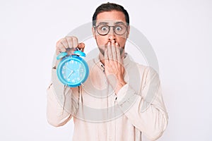 Young handsome man holding alarm clock covering mouth with hand, shocked and afraid for mistake