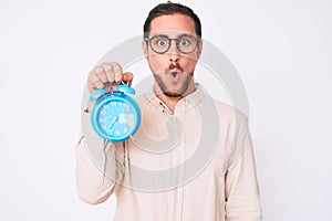 Young handsome man holding alarm clock scared and amazed with open mouth for surprise, disbelief face