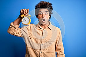 Young handsome man holding alarm clock standing over isolated blue background scared in shock with a surprise face, afraid and
