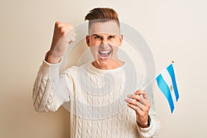 Young handsome man holding Argentinian Argentina flag over isolated white background annoyed and frustrated shouting with anger,