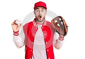 Young handsome man holding baseball gloves scared and amazed with open mouth for surprise, disbelief face