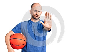 Young handsome man holding basketball ball with open hand doing stop sign with serious and confident expression, defense gesture