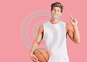 Young handsome man holding basketball ball surprised with an idea or question pointing finger with happy face, number one