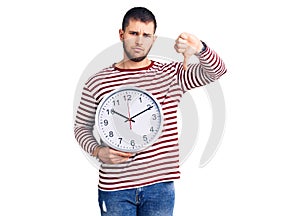 Young handsome man holding big clock with angry face, negative sign showing dislike with thumbs down, rejection concept