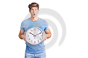 Young handsome man holding big clock scared and amazed with open mouth for surprise, disbelief face