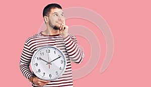 Young handsome man holding big clock serious face thinking about question with hand on chin, thoughtful about confusing idea