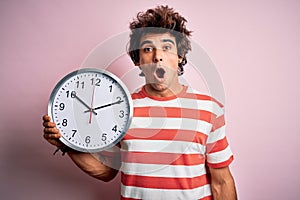 Young handsome man holding big clock standing over isolated pink background scared in shock with a surprise face, afraid and