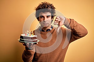 Young handsome man holding birthday cake standing over isolated yellow background with angry face, negative sign showing dislike