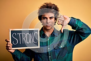Young handsome man holding blackboard with strong word over isolated yellow background with angry face, negative sign showing
