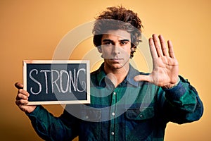 Young handsome man holding blackboard with strong word over isolated yellow background with open hand doing stop sign with serious