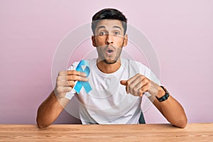 Young handsome man holding blue ribbon scared and amazed with open mouth for surprise, disbelief face