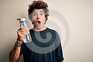 Young handsome man holding bottle of water standing over isolated white background scared in shock with a surprise face, afraid