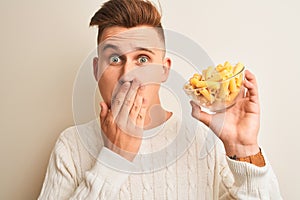 Young handsome man holding bowl with dry pasta standing over isolated white background cover mouth with hand shocked with shame