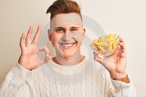 Young handsome man holding bowl with dry pasta standing over isolated white background doing ok sign with fingers, excellent