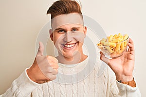 Young handsome man holding bowl with dry pasta standing over isolated white background happy with big smile doing ok sign, thumb