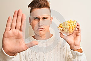 Young handsome man holding bowl with dry pasta standing over isolated white background with open hand doing stop sign with serious