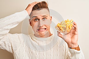 Young handsome man holding bowl with dry pasta standing over isolated white background stressed with hand on head, shocked with