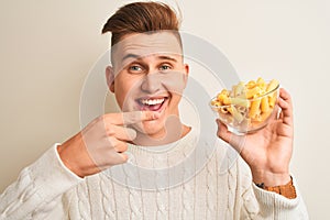 Young handsome man holding bowl with dry pasta standing over isolated white background very happy pointing with hand and finger