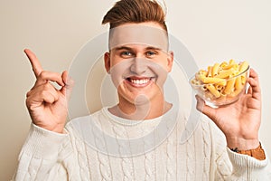 Young handsome man holding bowl with dry pasta standing over isolated white background very happy pointing with hand and finger to