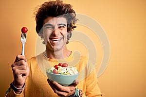 Young handsome man holding bowl with salad standing over isolated yellow background with a happy face standing and smiling with a