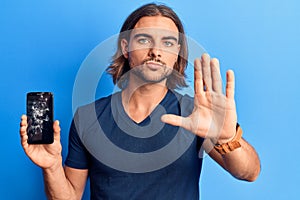 Young handsome man holding broken smartphone showing cracked screen with open hand doing stop sign with serious and confident