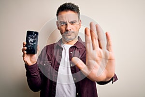 Young handsome man holding broken smartphone showing craked screen with open hand doing stop sign with serious and confident