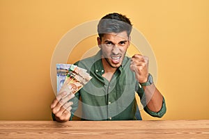 Young handsome man holding canadian dollars angry and mad raising fist frustrated and furious while shouting with anger