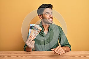 Young handsome man holding canadian dollars looking away to side with smile on face, natural expression