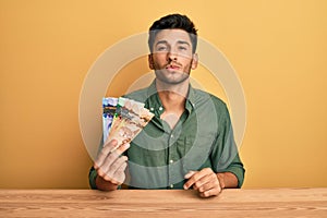 Young handsome man holding canadian dollars looking at the camera blowing a kiss on air being lovely and sexy