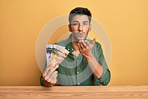 Young handsome man holding canadian dollars looking at the camera blowing a kiss with hand on air being lovely and sexy