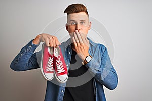 Young handsome man holding casual sneakers standing over isolated white background cover mouth with hand shocked with shame for