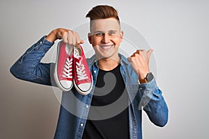 Young handsome man holding casual sneakers standing over isolated white background pointing and showing with thumb up to the side