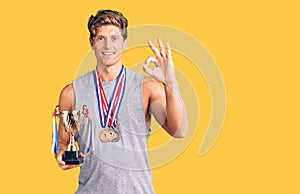 Young handsome man holding champion trophy wearing medals doing ok sign with fingers, smiling friendly gesturing excellent symbol