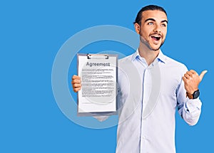 Young handsome man holding clipboard with agreement document pointing thumb up to the side smiling happy with open mouth