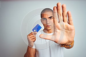 Young handsome man holding credit card over white isolated background with open hand doing stop sign with serious and confident