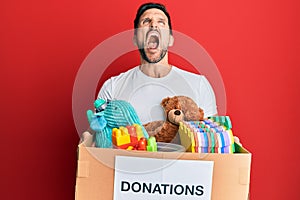 Young handsome man holding donation box with toys angry and mad screaming frustrated and furious, shouting with anger looking up