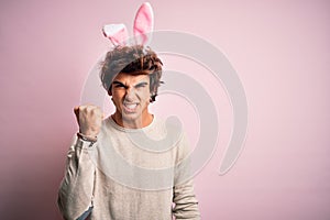 Young handsome man holding easter rabbit ears standing over isolated pink background angry and mad raising fist frustrated and