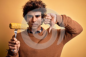 Young handsome man holding fork with cob corn standing over isolated yellow background with angry face, negative sign showing