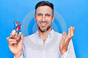 Young handsome man holding heart organ with veins and arteries over blule background celebrating achievement with happy smile and